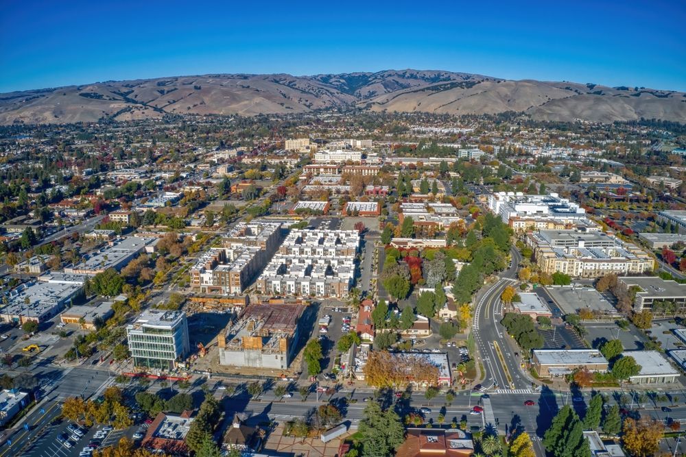 Aerial view of a city with buildings, roads, and hills in the background under a blue sky.