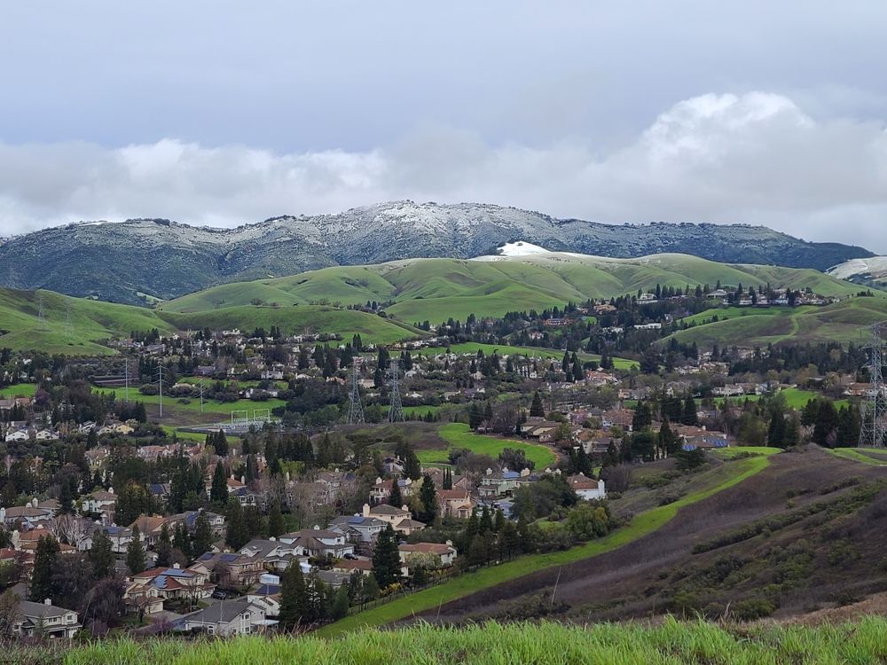 Green hills and suburban homes under a cloudy sky. Snow on the distant mountain peaks.
