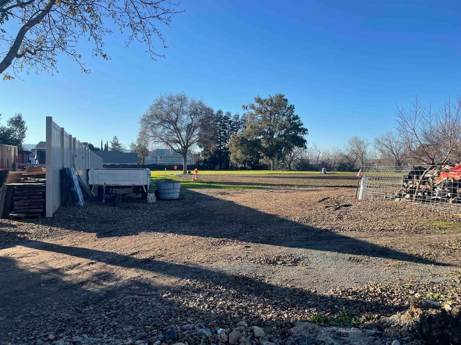 Gravel-covered lot with shadows cast by trees and structures.  A few trees, debris, and clear blue sky are visible.