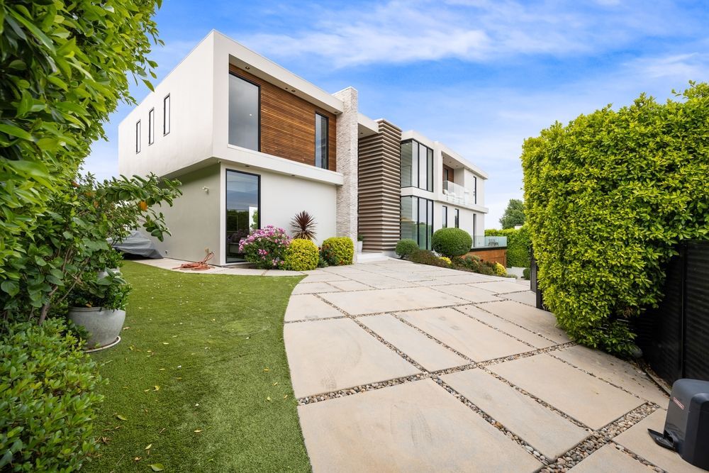Modern white house with large windows, surrounded by green landscaping and a concrete driveway.