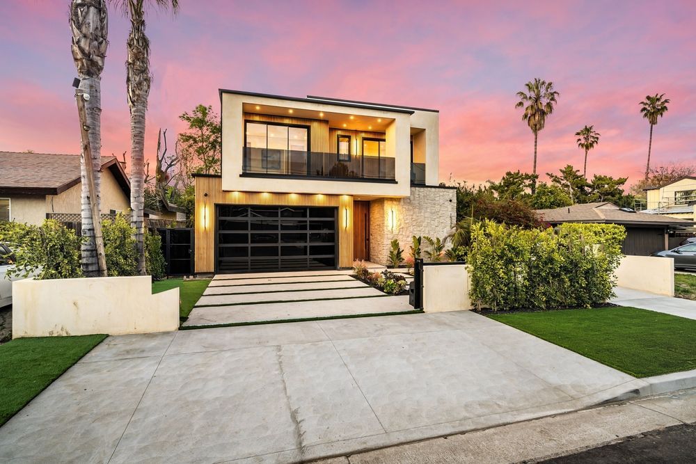 Modern two-story house with a glass garage door and balcony against a sunset sky.