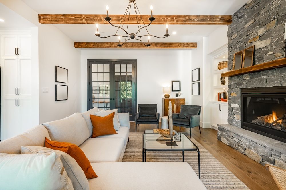 Cozy living room with a stone fireplace, cream sofa, and dark French doors. Wooden beams and a chandelier.