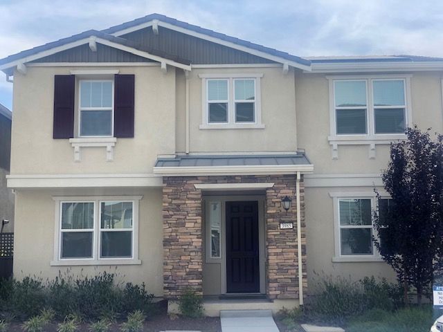 Two-story beige house with stone accents around the door. Red shutters on one window.