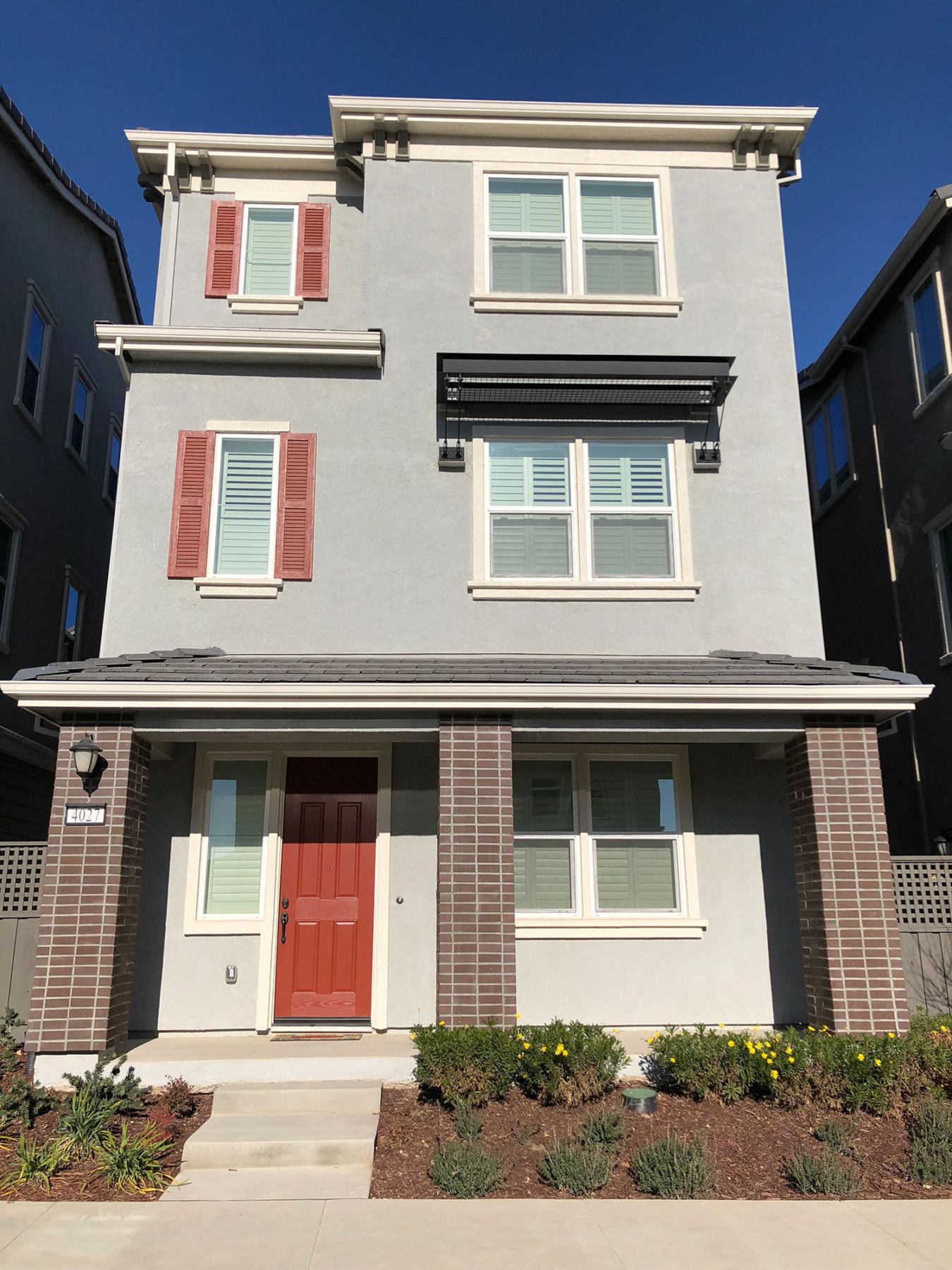 Three-story house with gray stucco, red door, shutters, and awnings. Brick columns frame the porch.