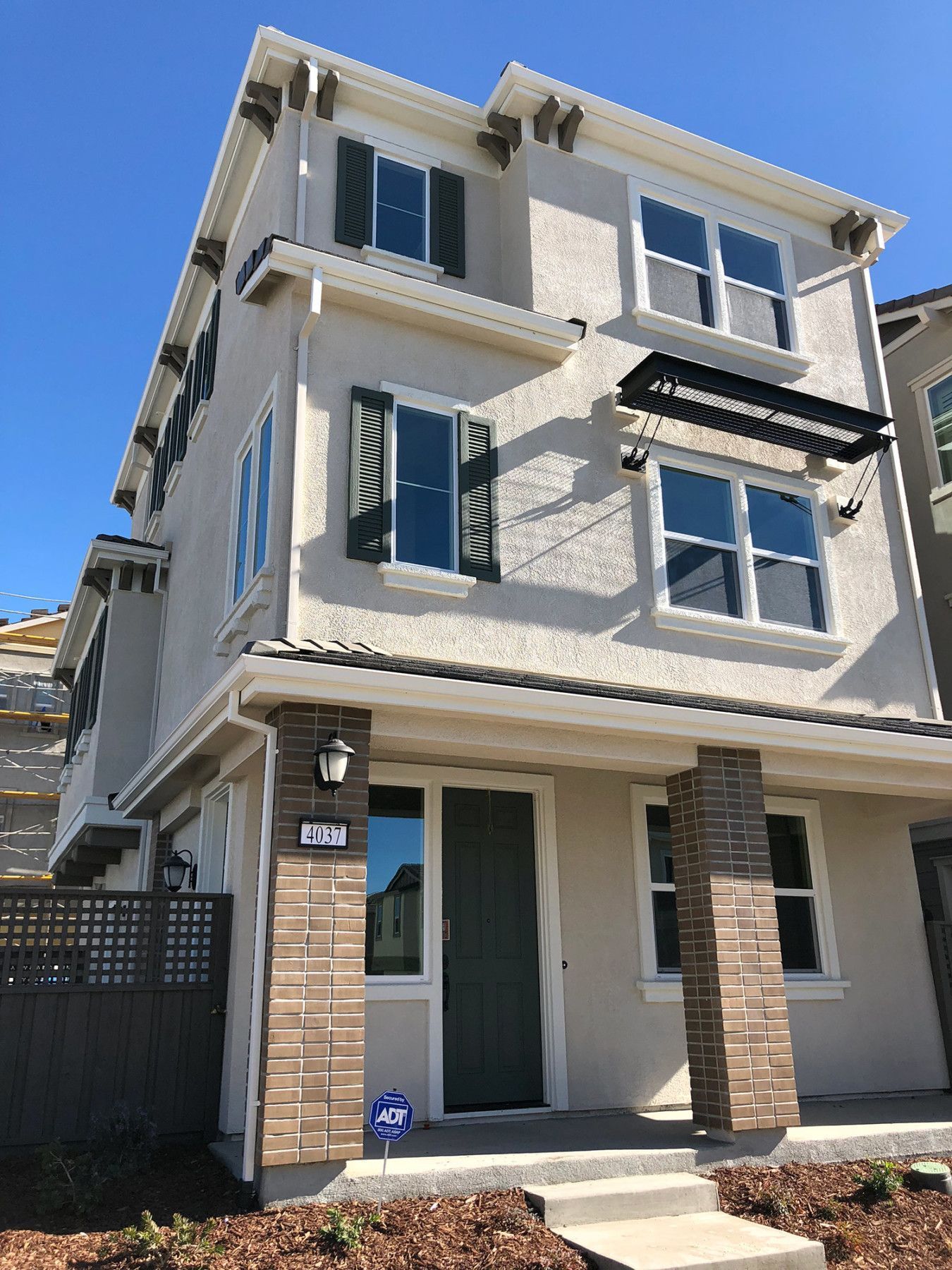 Three-story beige townhome with green shutters, brown accents, and a black awning under a clear blue sky.