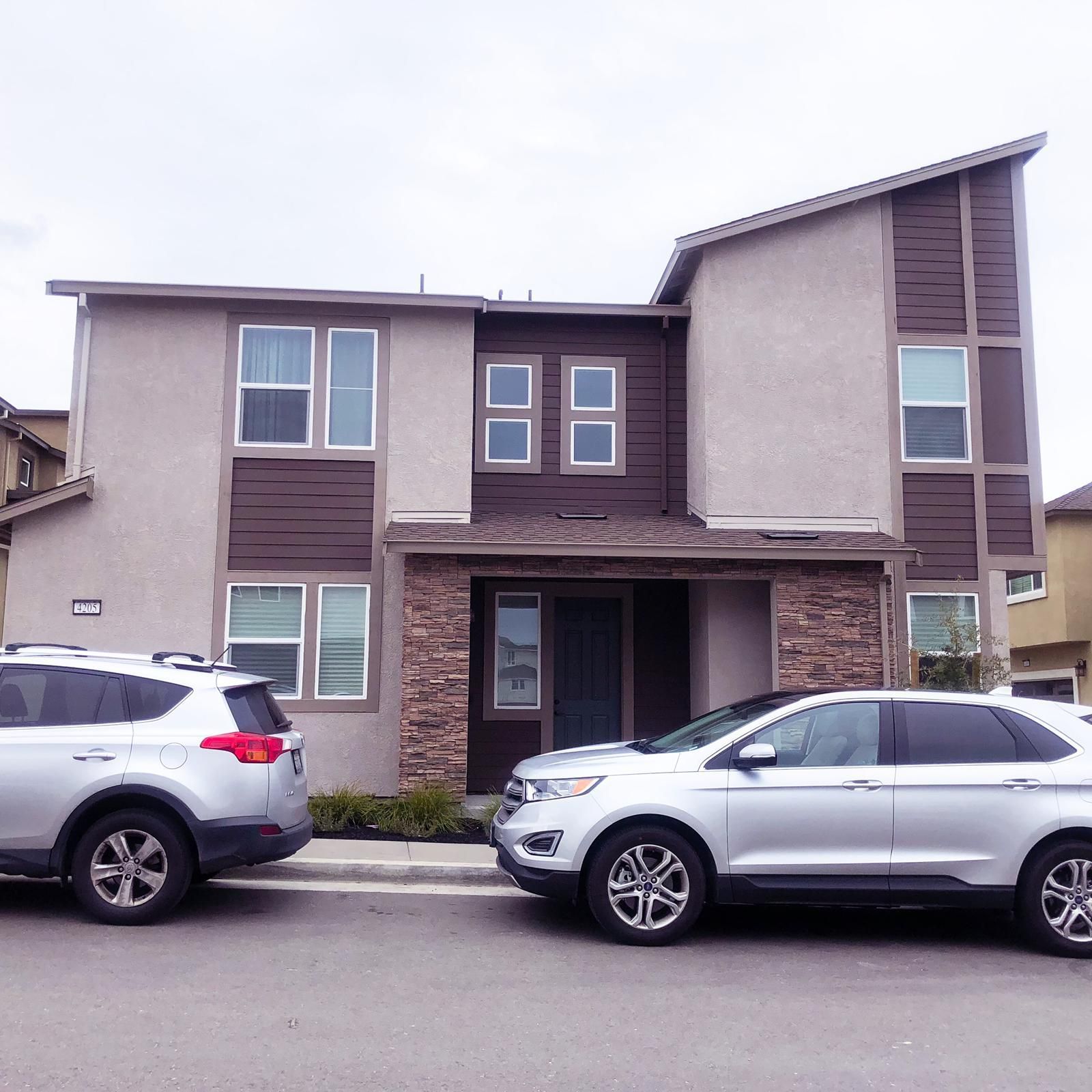 Two-story house with tan stucco and brown trim, two silver SUVs parked in front.