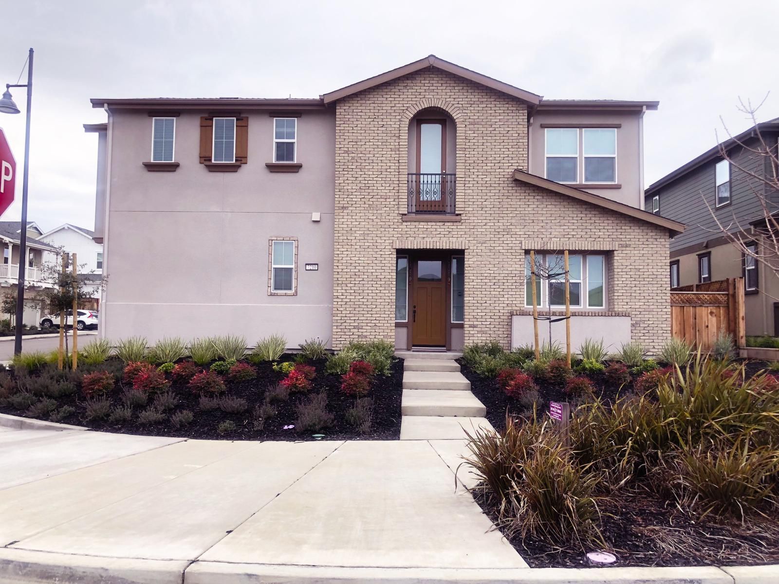 Two-story house with tan brick facade, small porch, and manicured landscaping, against cloudy sky.