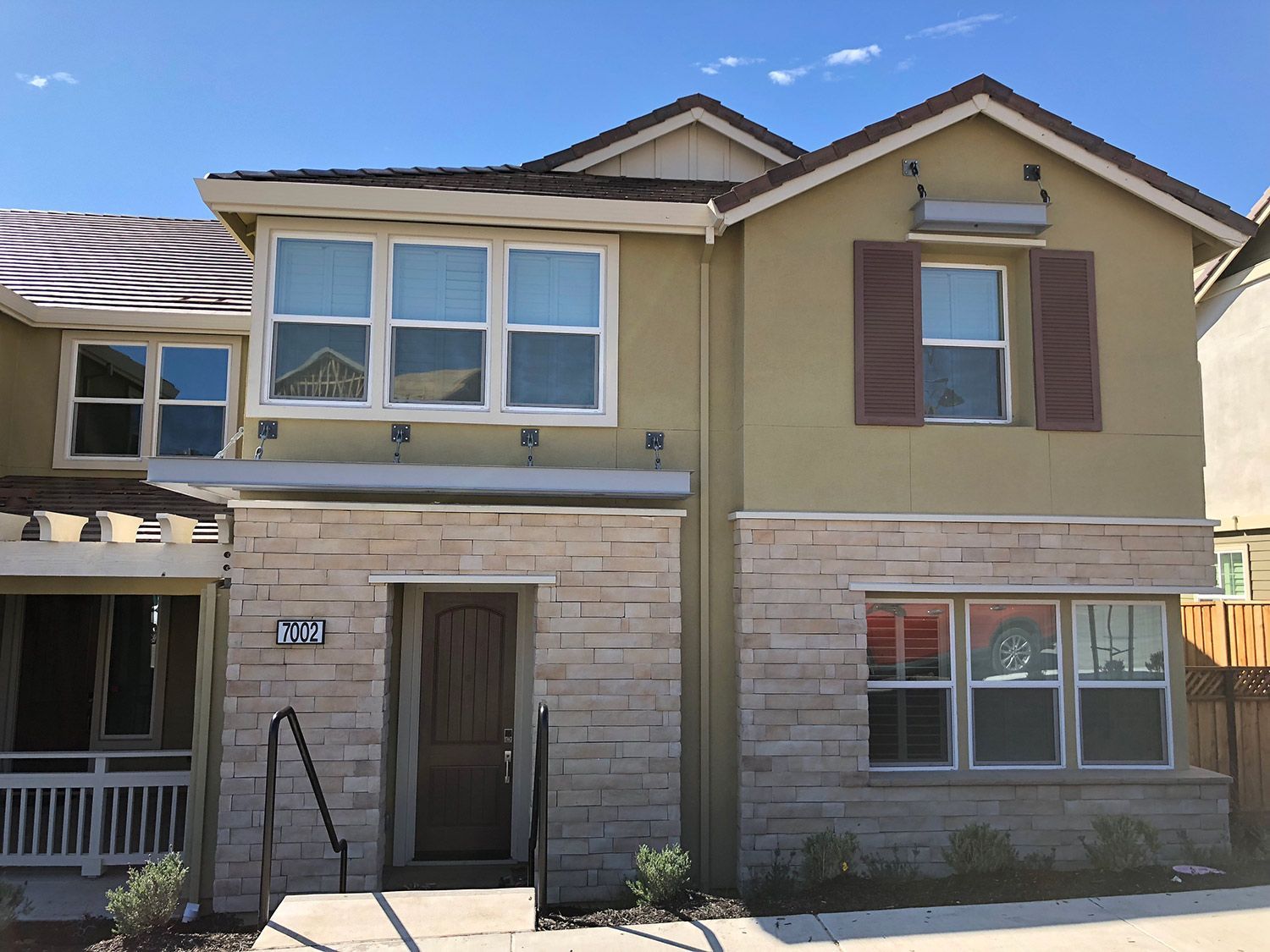 Two-story beige house with stone accents, brown shutters, and multiple windows on a sunny day.