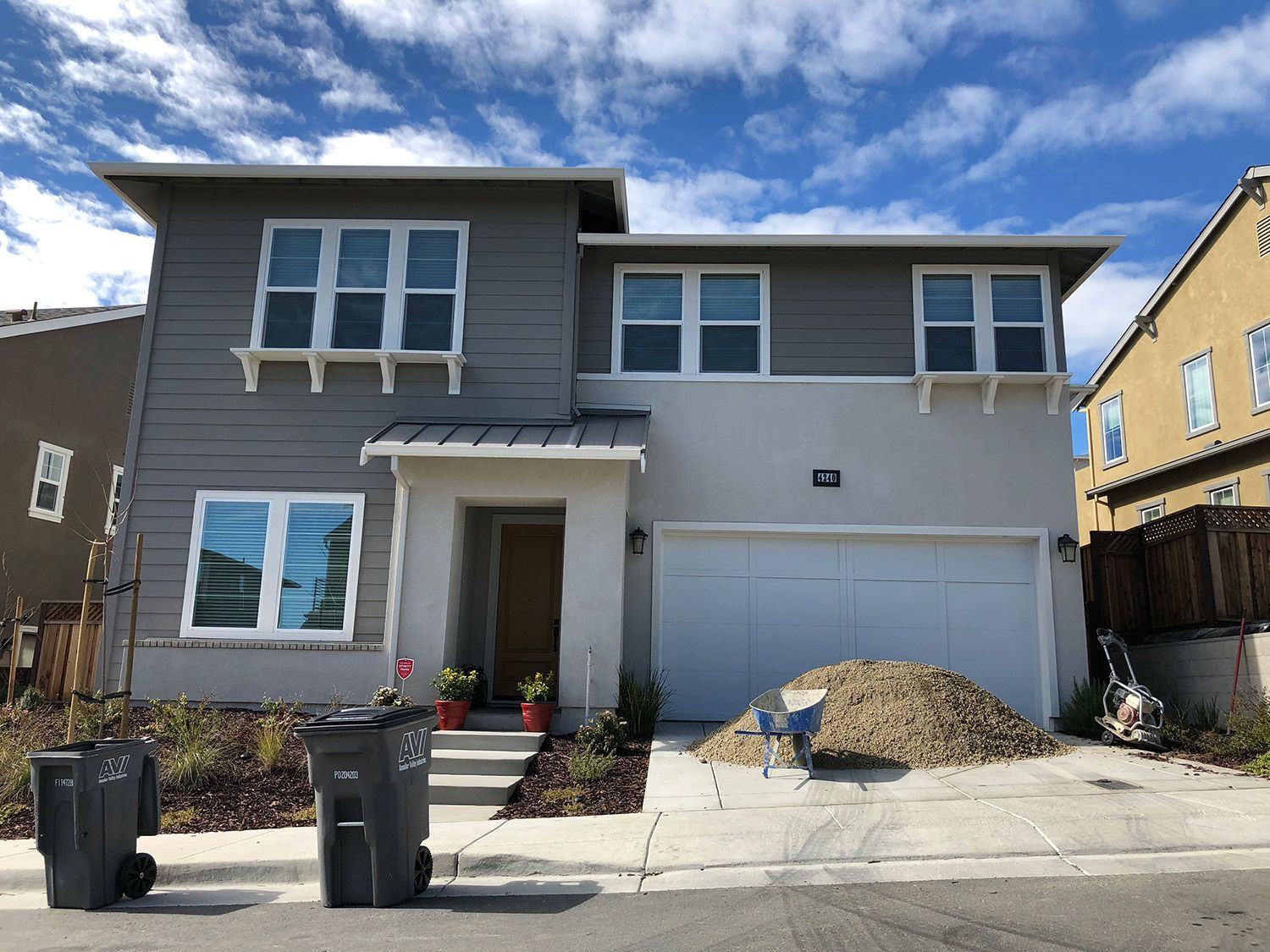 Two-story house with gray and tan siding, two trash cans in the foreground, and a pile of gravel in front of the garage.