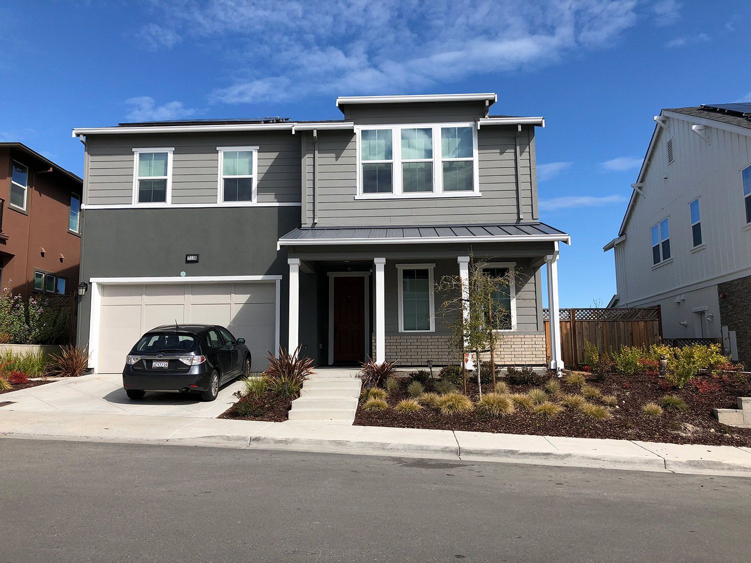 Two-story gray house with white trim, a red door, and a car parked in the driveway on a sunny day.
