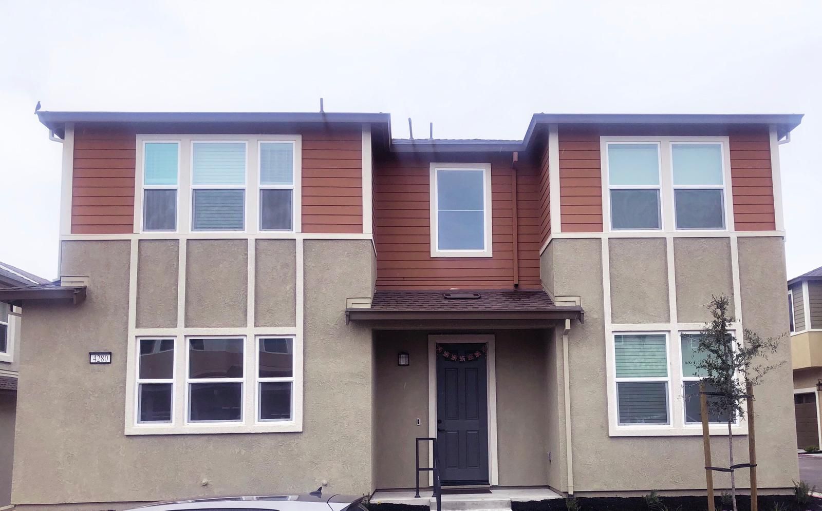 Two-story tan and brown house with multiple windows, dark front door, and a cloudy sky.