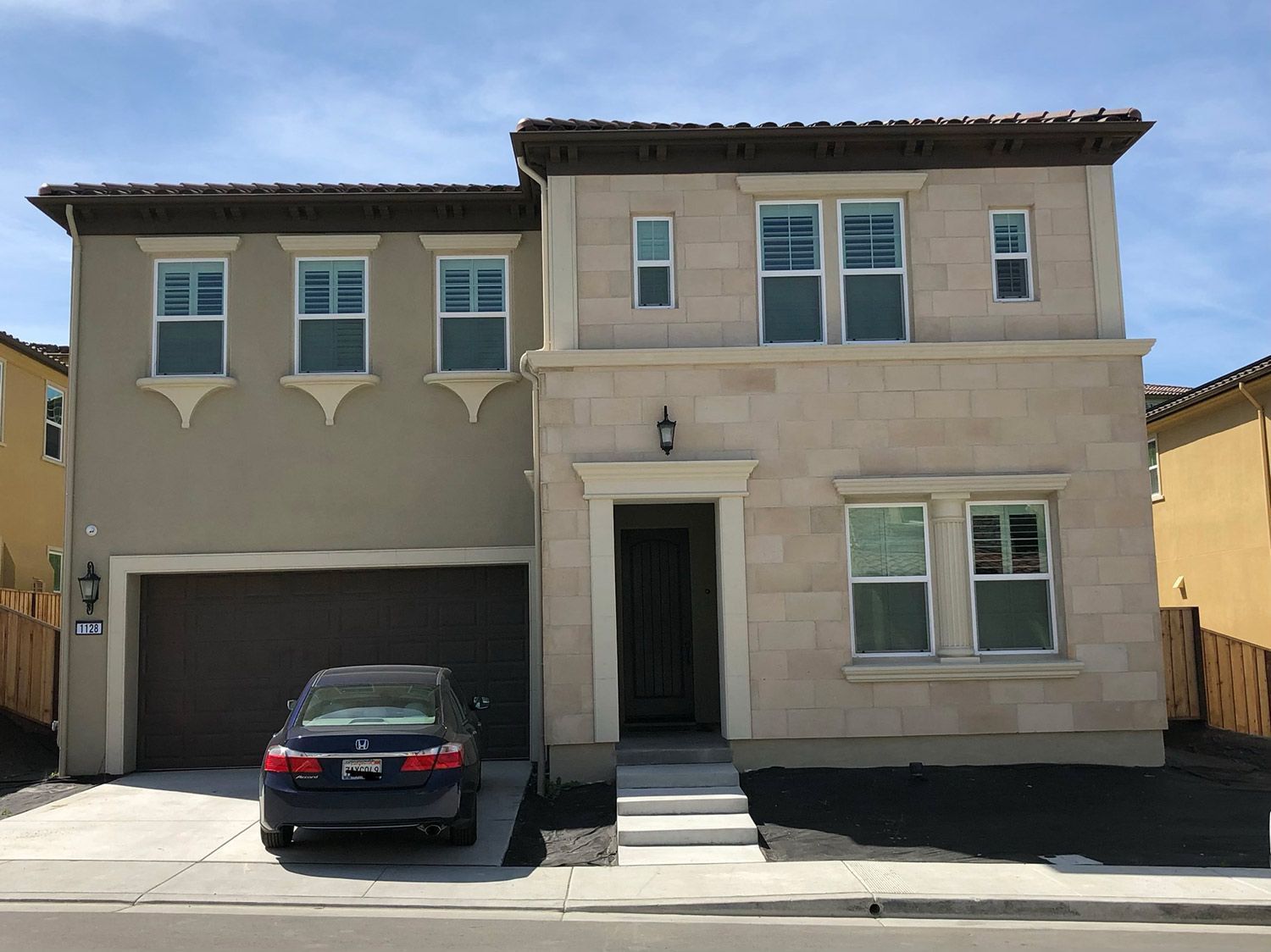 Two-story beige house with a dark car in the driveway and a brown garage door under a blue sky.