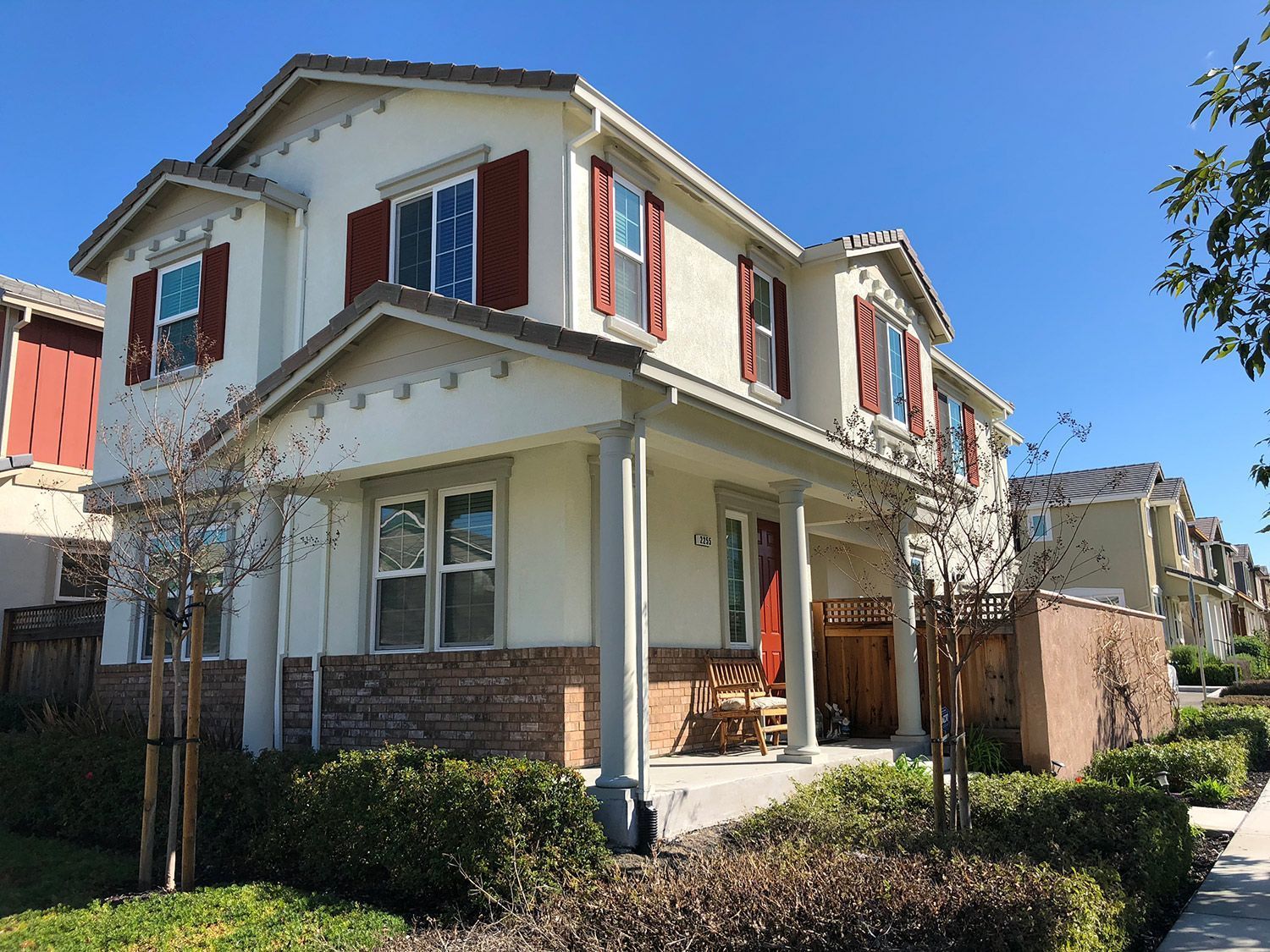 Two-story beige house with red shutters and front porch; blue sky.