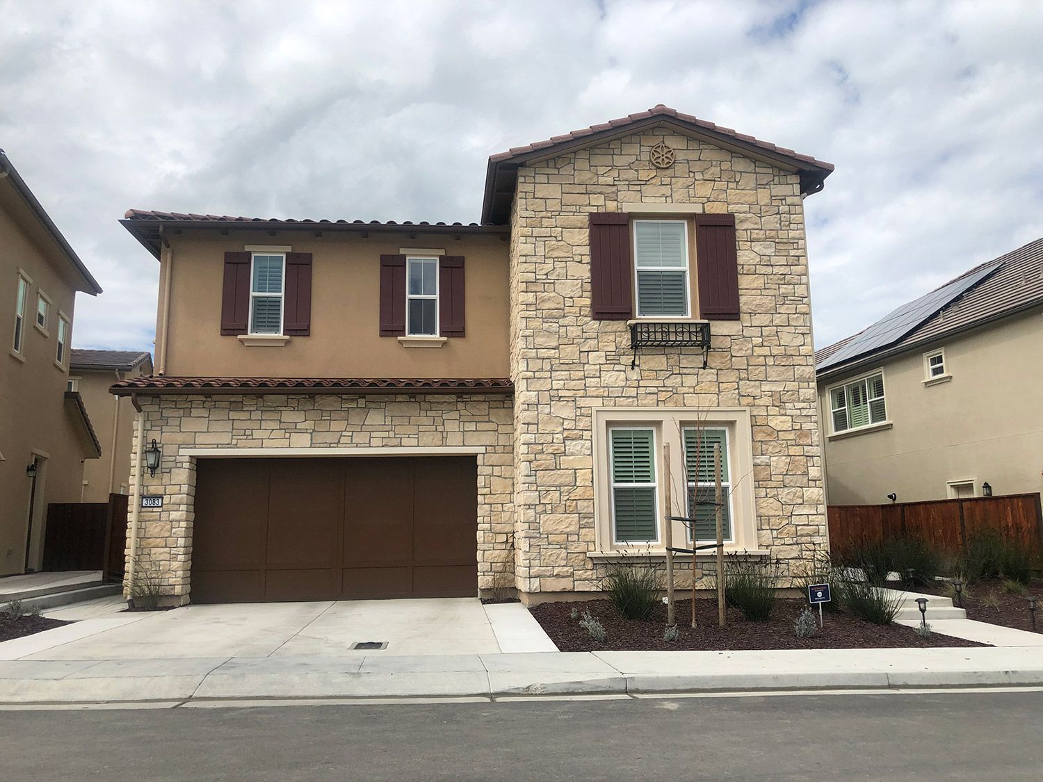Two-story house with stone facade, brown garage door, and tan stucco siding under cloudy sky.