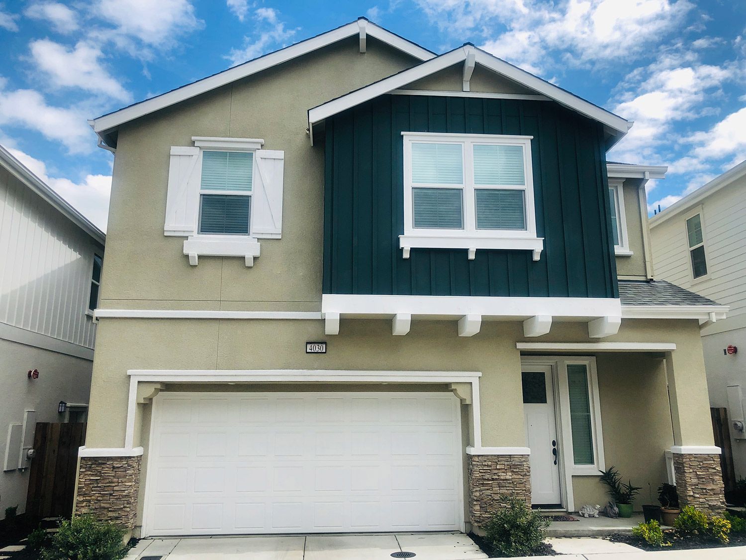Two-story house with tan stucco, white trim, and a dark green accent. A white garage door and blue sky are visible.