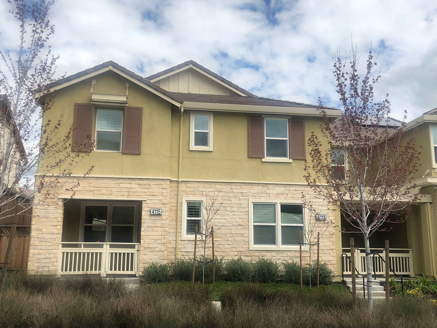 Two-story beige townhome with stone facade, shuttered windows, and porch, under cloudy sky.
