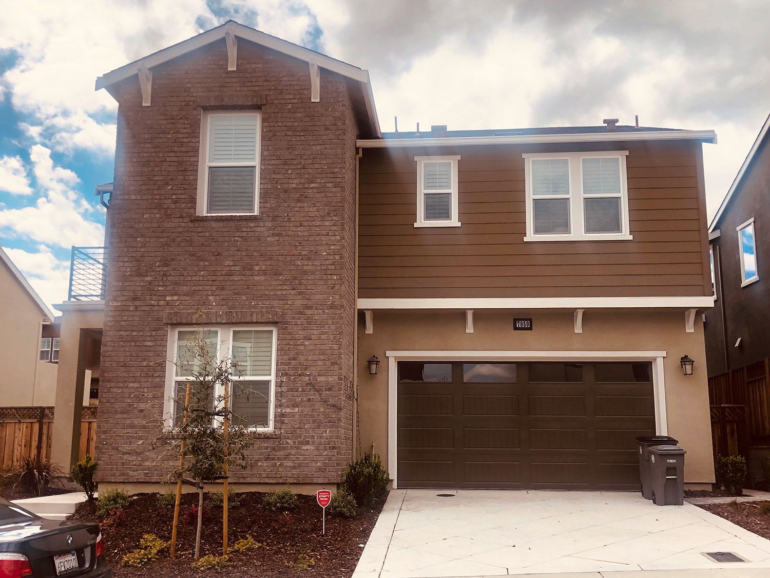 Two-story house with brown siding and garage. Brick-textured facade on the left side. Cloudy sky.