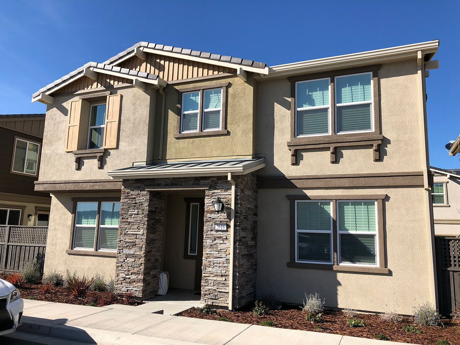 Two-story house with stone accents, tan siding, and brown trim against a clear blue sky.