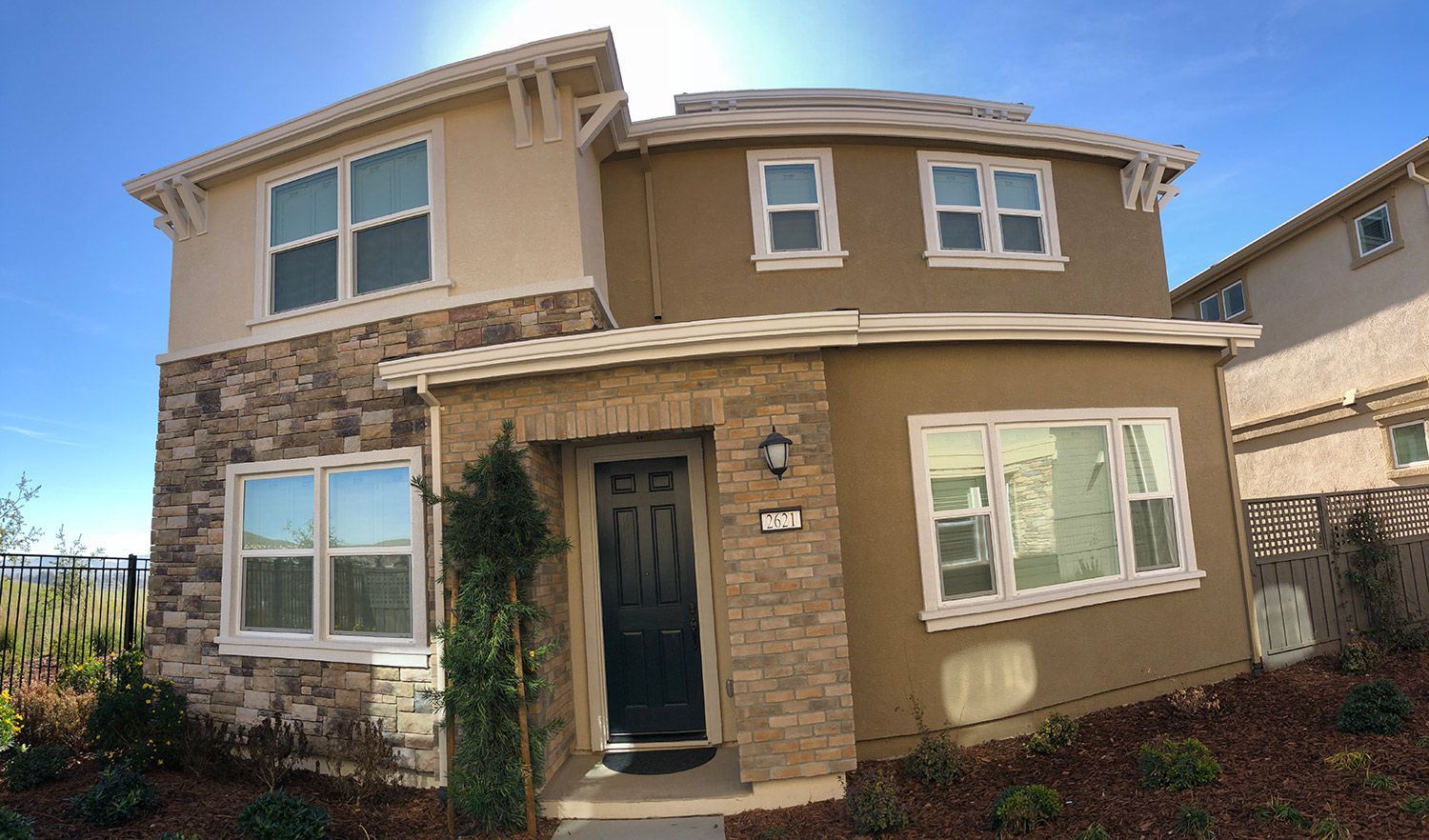 Two-story house with stone and tan stucco exterior, windows, and a dark front door under a sunny sky.