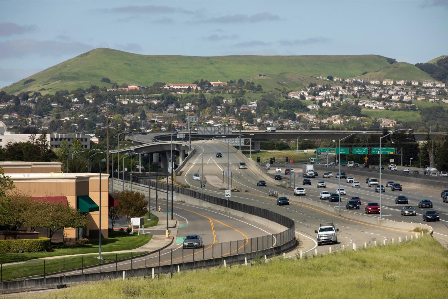 Highway with traffic curves toward a hillside residential area under a blue sky.