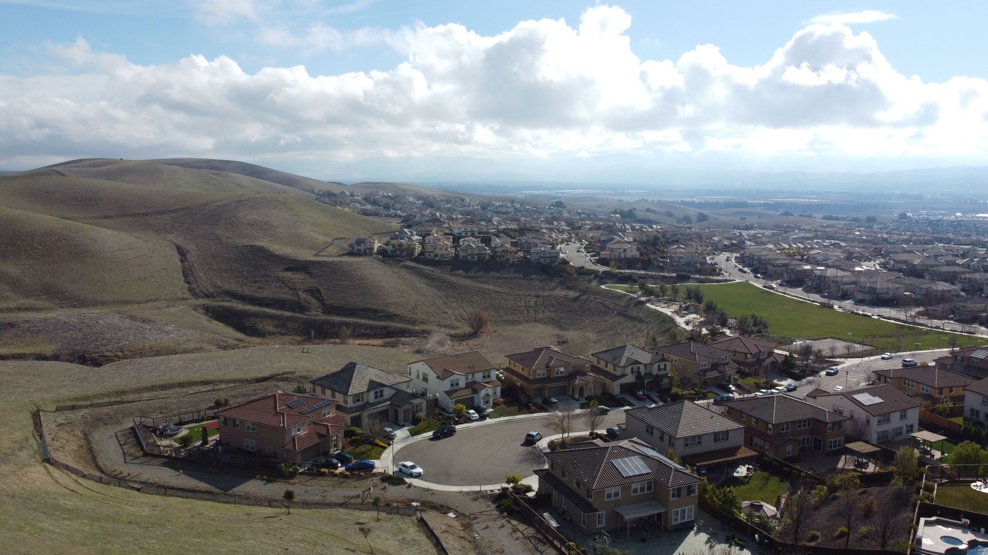 Panoramic view of a hillside town with houses, a green valley, and cloudy sky.