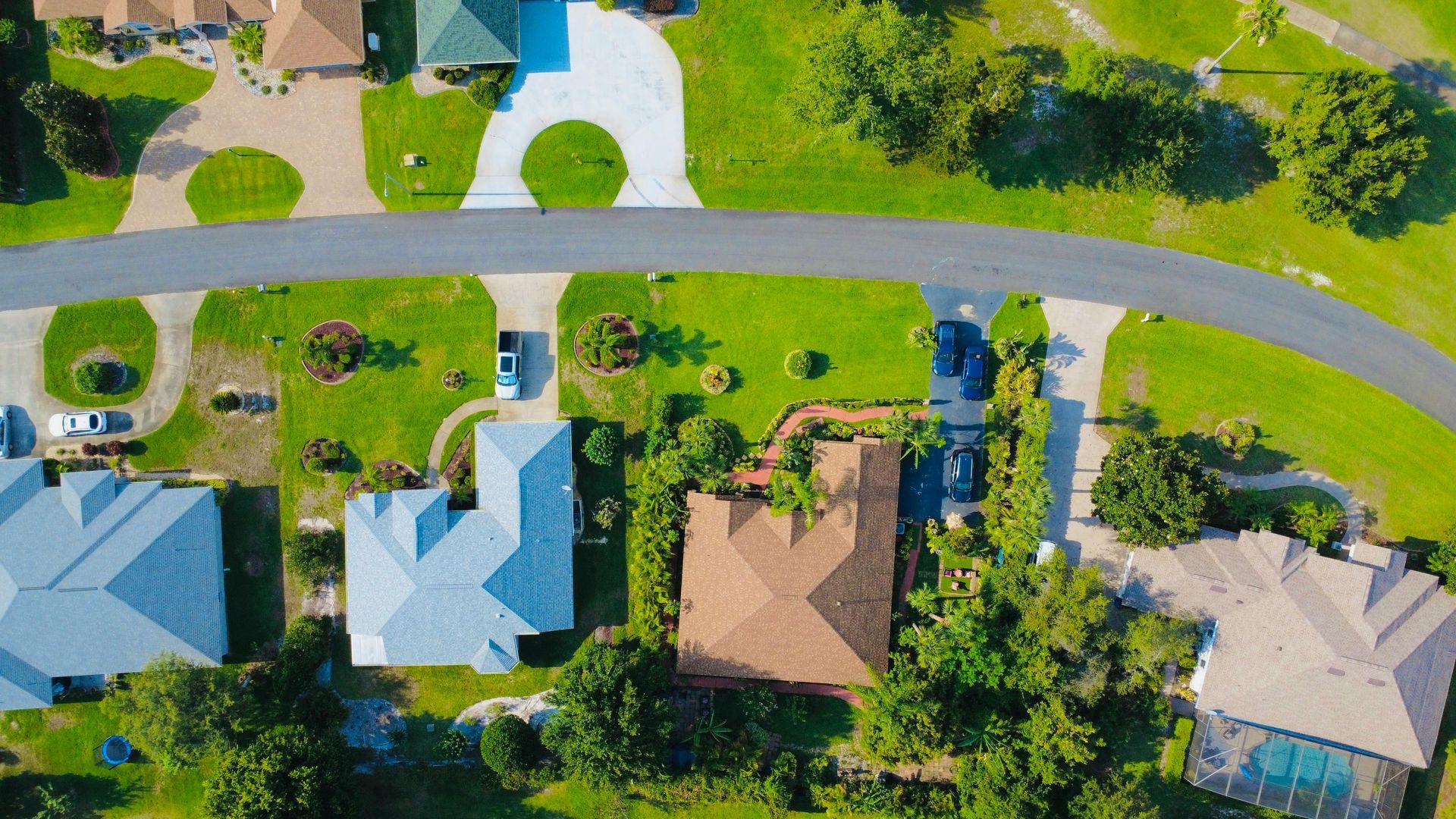 Aerial view of houses with blue and brown roofs, a curved road, and green lawns.