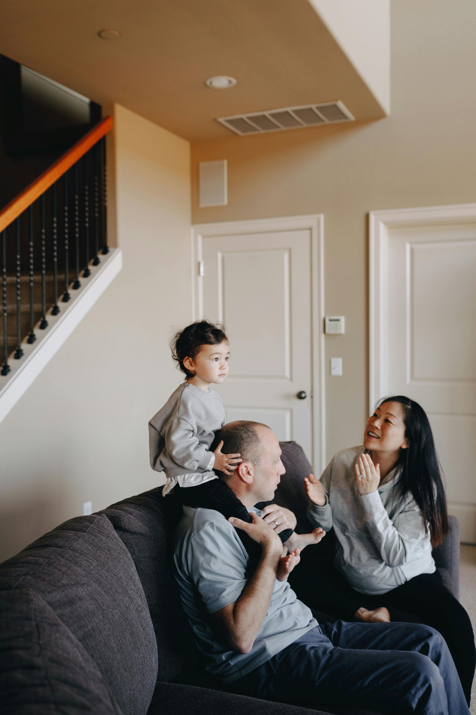 A child sits on a person’s shoulders on a grey couch, while another person sits nearby, all in a home interior.