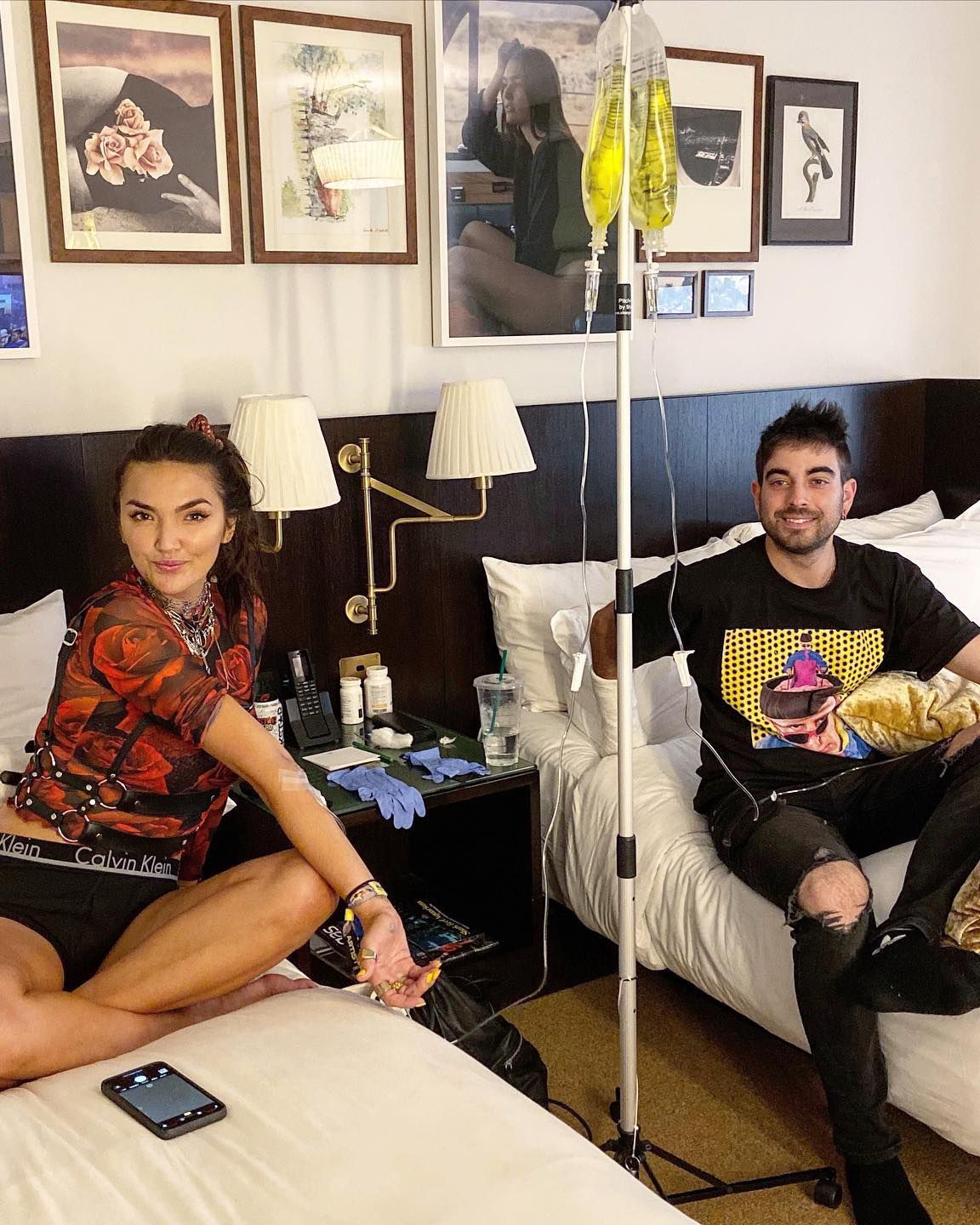 Two people sitting on a bed in a hotel room, with a mirror, lamp, and framed art on the wall.