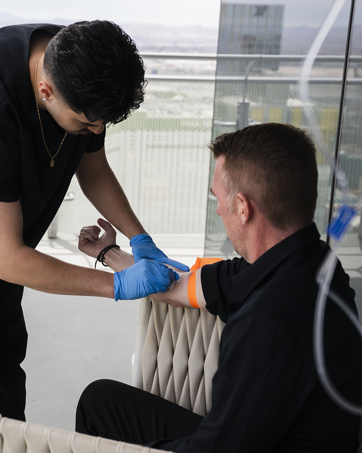 Healthcare worker drawing blood from a seated patient by a window, wearing blue gloves and an orange tourniquet.