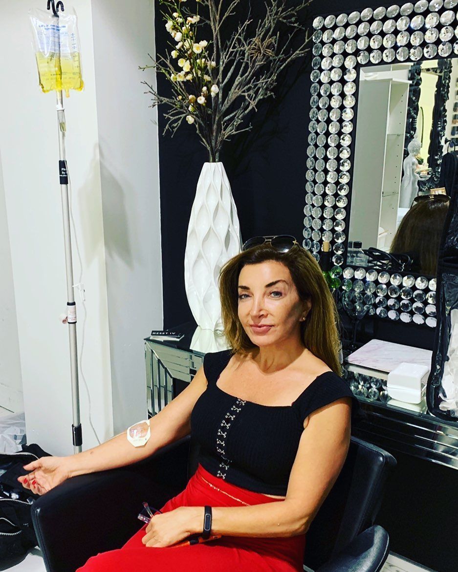 Woman seated in a salon chair, wearing a black top and red skirt, beside a mirror and white vase of branches
