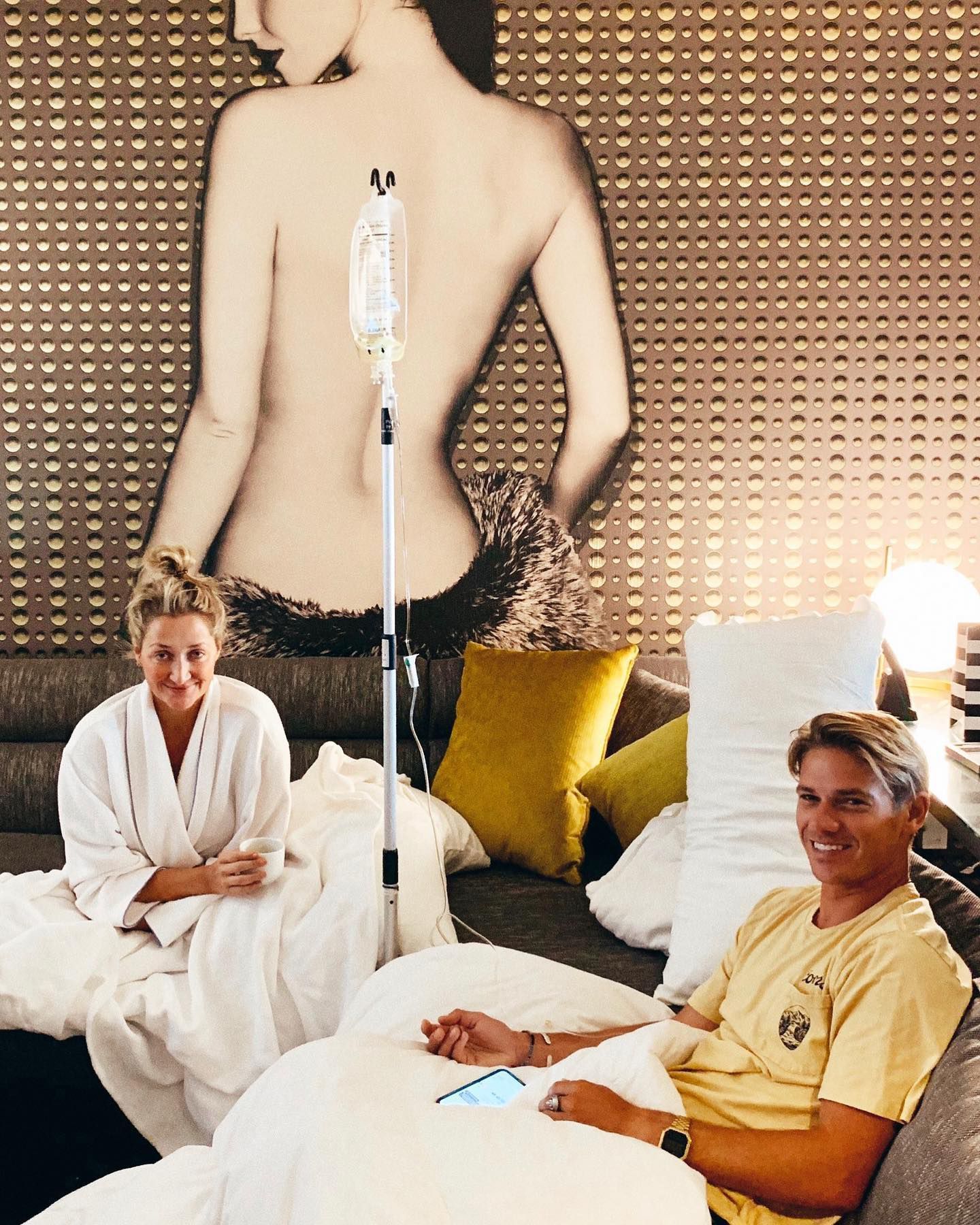 Two people in white robes relax on a bed in a spa room with an IV stand and large wall art.