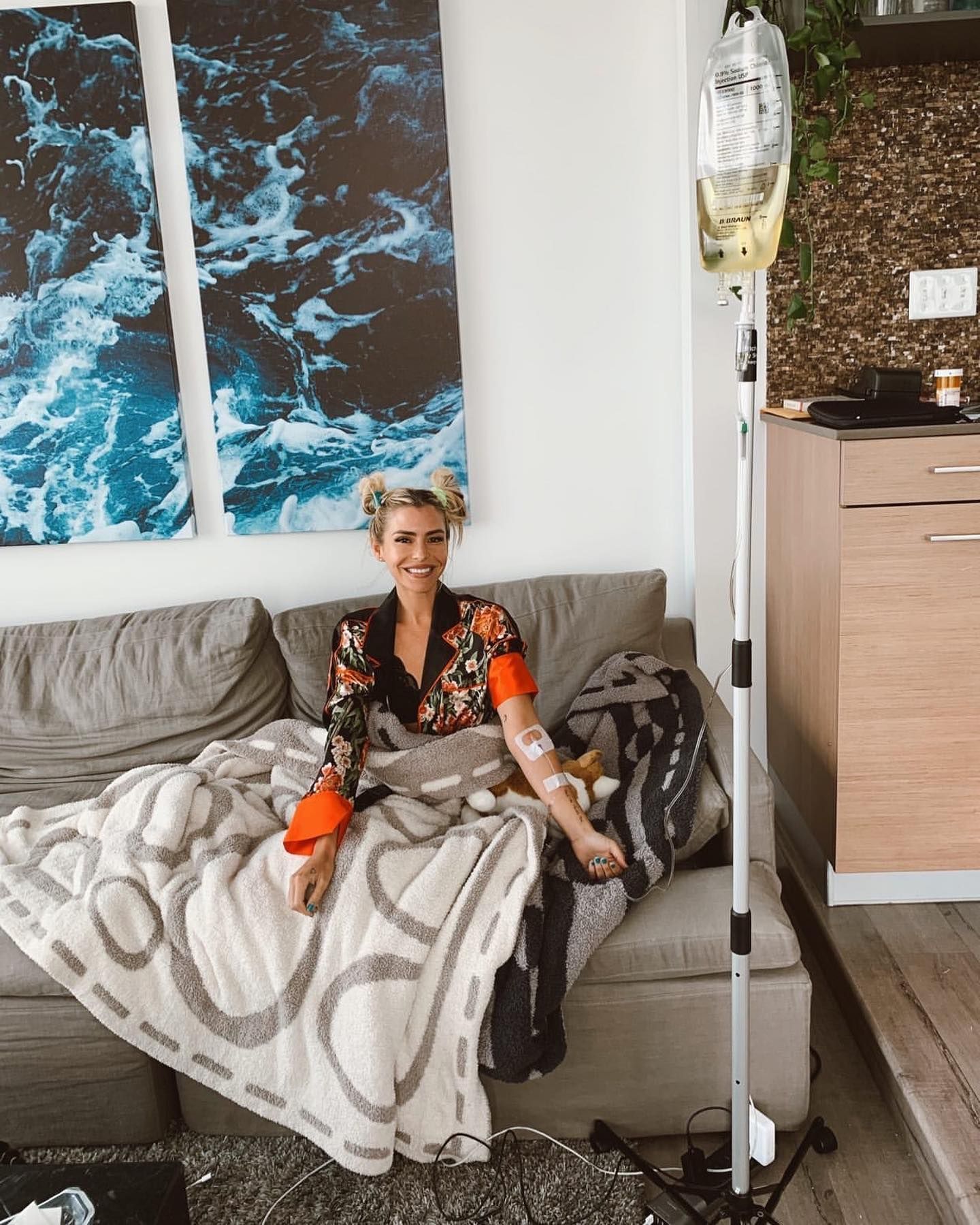 Person seated on a gray couch with a blanket, in a bright living room with ocean artwork on the wall.