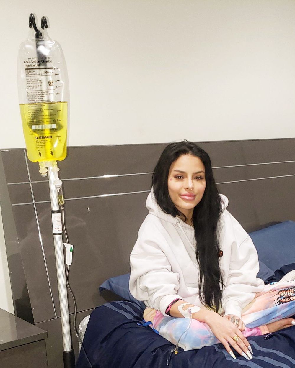 Woman seated in a hospital bed beside an IV stand, smiling at the camera.
