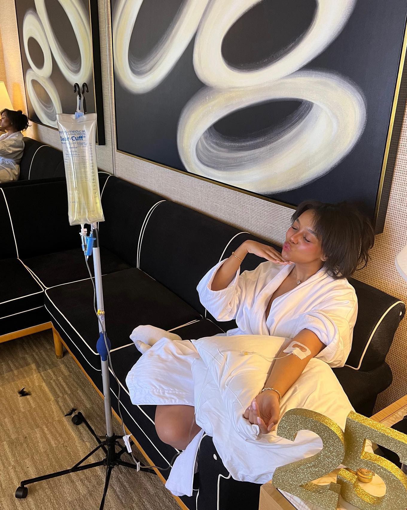 Woman sitting on a couch beside a medical IV pole in a sparse living room