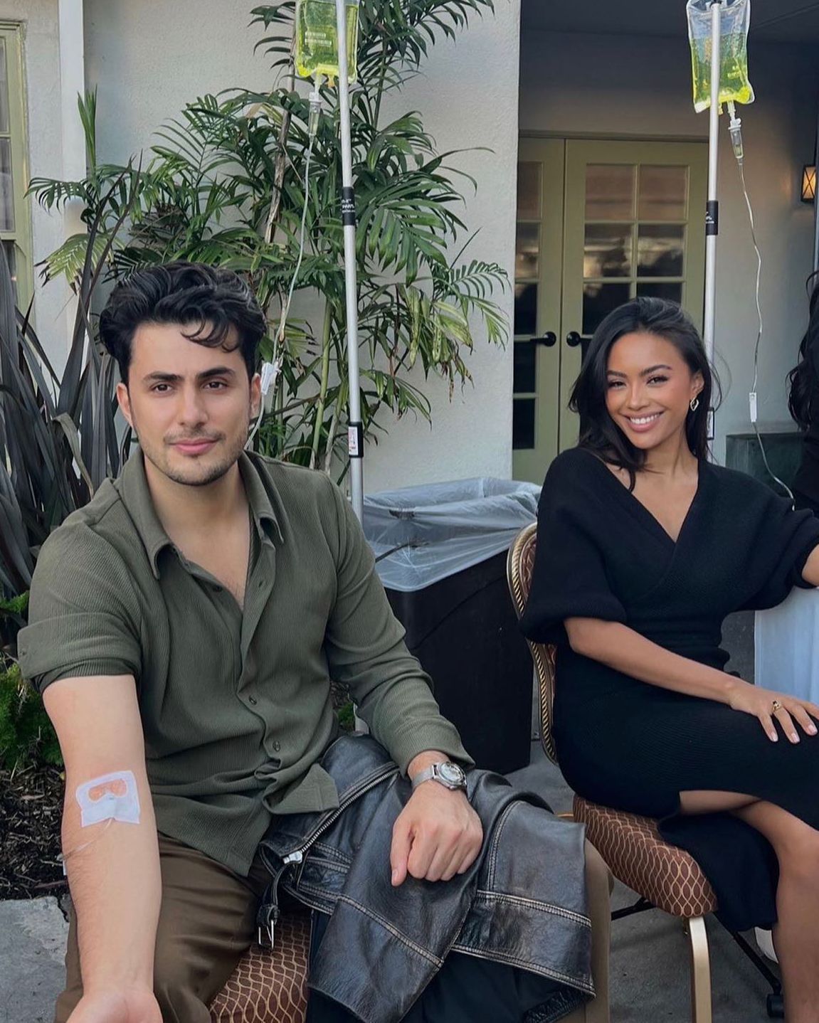 Two people seated outdoors on a patio, smiling, with potted plants and hanging decor behind them
