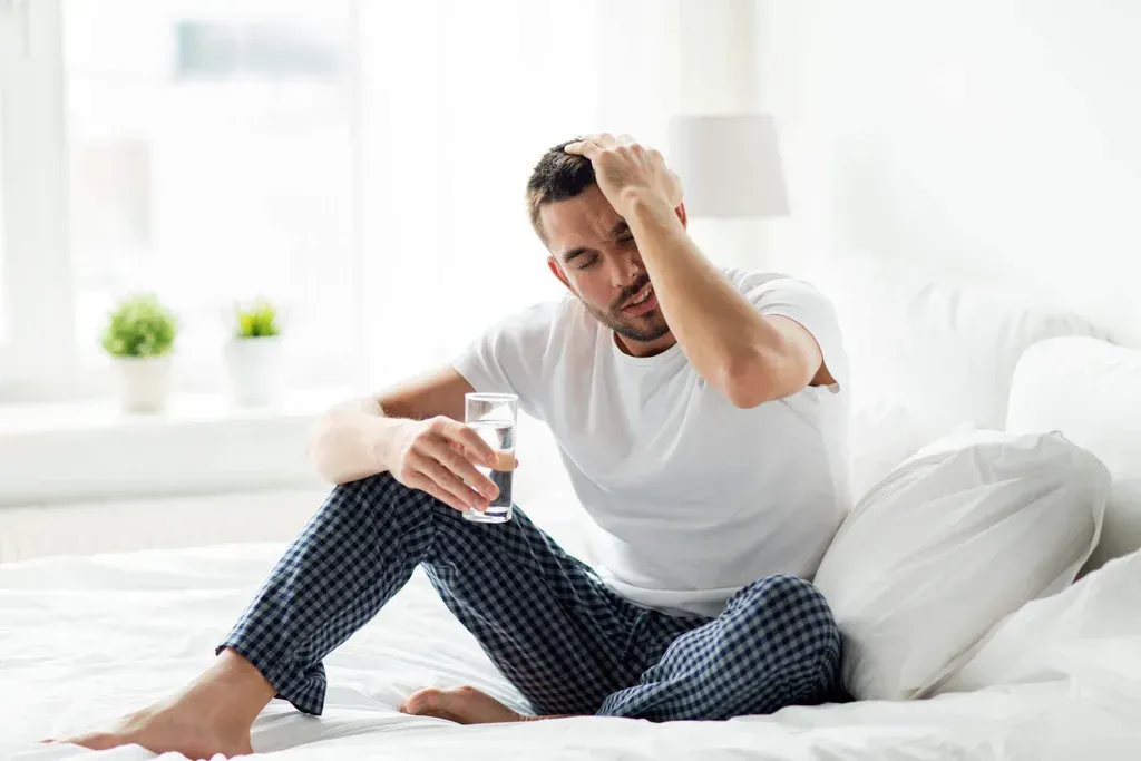 Man sitting on a bed, holding his head and a glass of water, looking unwell in a bright bedroom