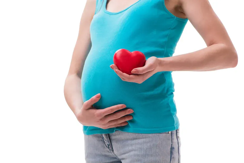 Pregnant person holding a red heart over their belly, wearing a turquoise tank top and jeans