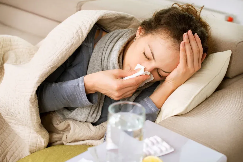 Sick person in bed holding a tissue and covering their face, with medicine and water on a bedside table