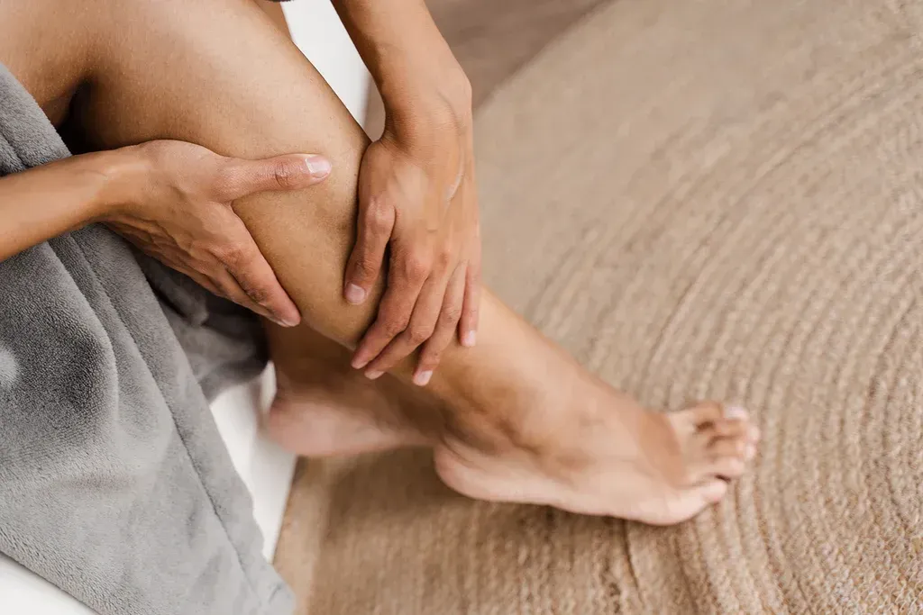 Person applying lotion to a leg on a wooden floor, with a gray towel nearby