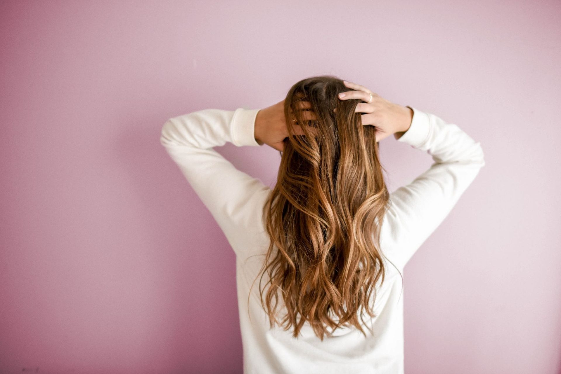 Person seen from behind, hands in long brown hair, against a pink wall