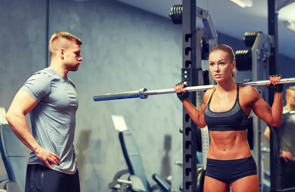 Man and woman in a gym, one spotting while the other holds a barbell on her shoulders