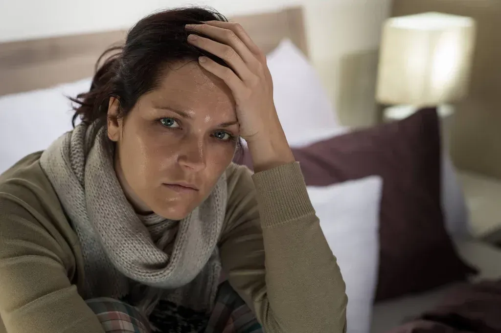Woman sitting on a bed, resting her head on her hand with a worried expression in a softly lit bedroom