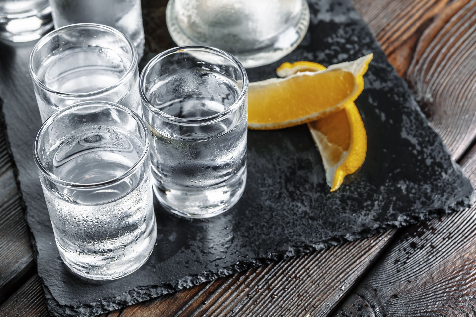 Four shot glasses of clear liquid on a slate tray with orange slices on a wooden table
