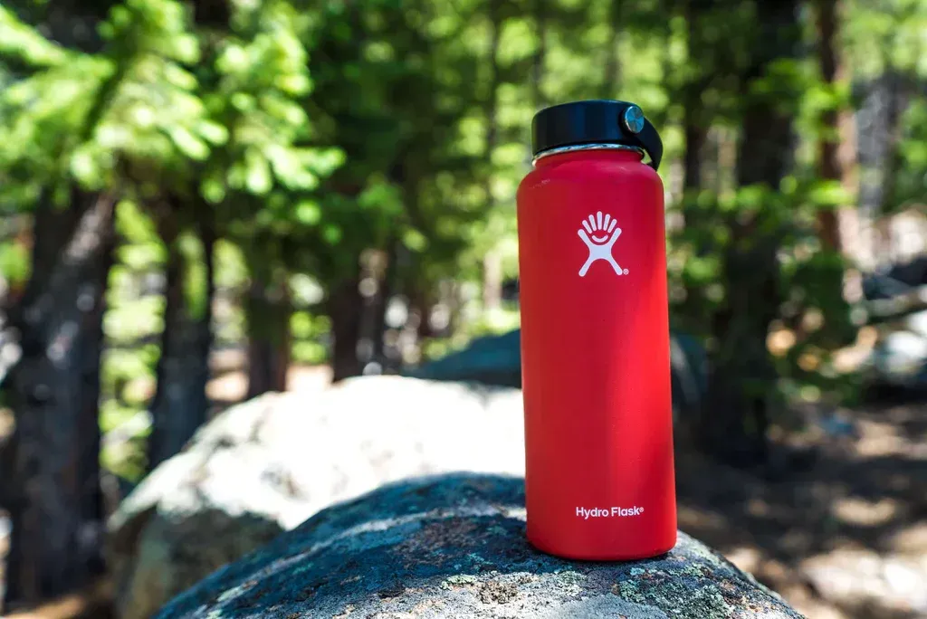 Red water bottle with a black cap on a rock in a forest, with trees blurred in the background