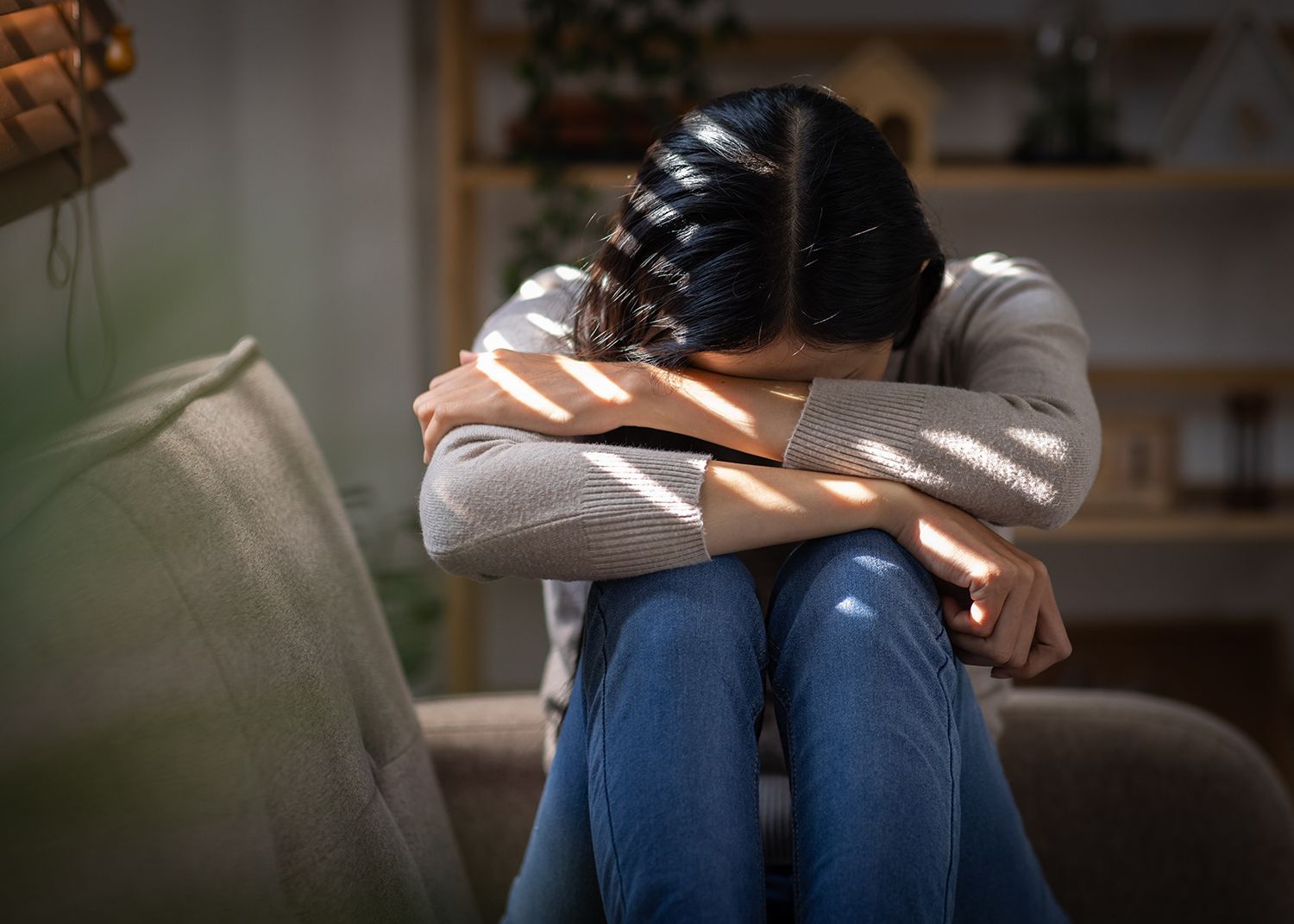 Person sitting curled up on a couch with head down and arms wrapped around knees in a dim room