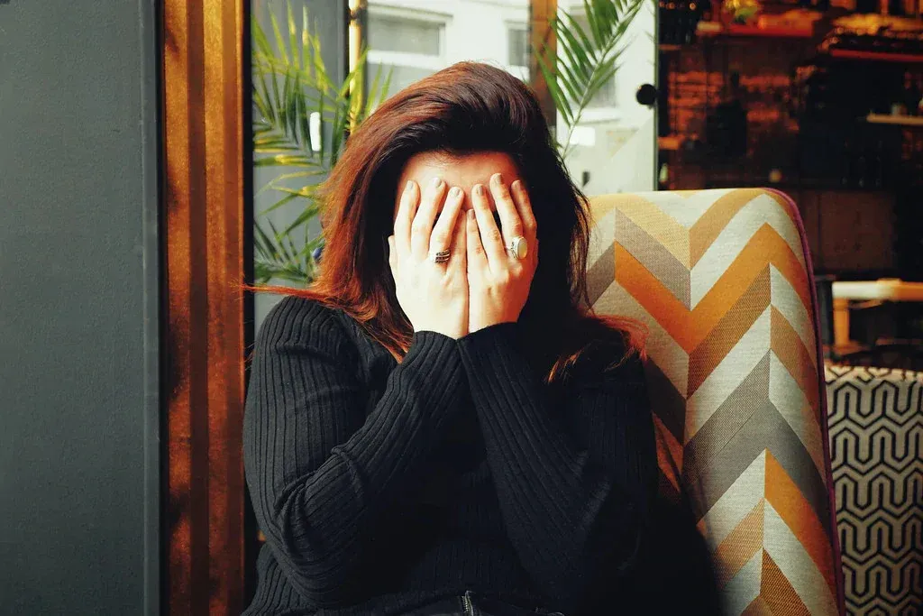 Person in a black sweater covers their face while sitting by a window in a cozy indoor setting.