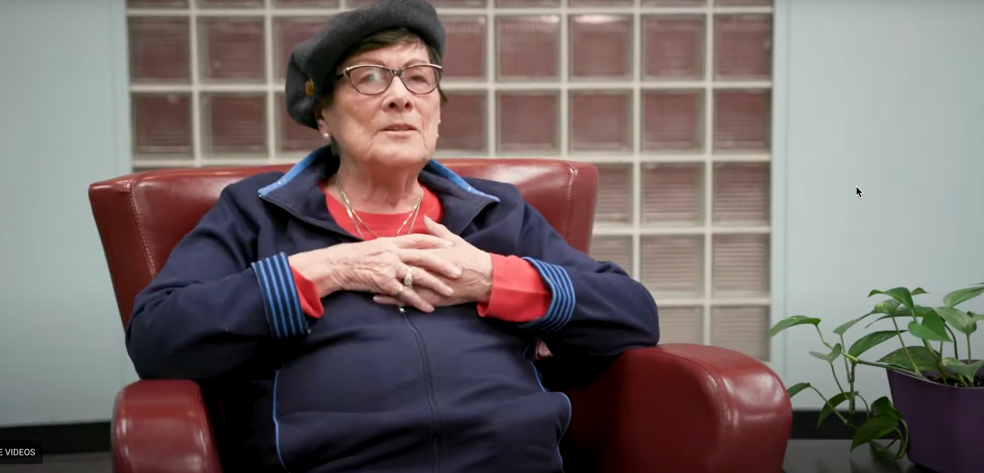 Une femme âgée est assise sur une chaise rouge, les mains sur la poitrine.
