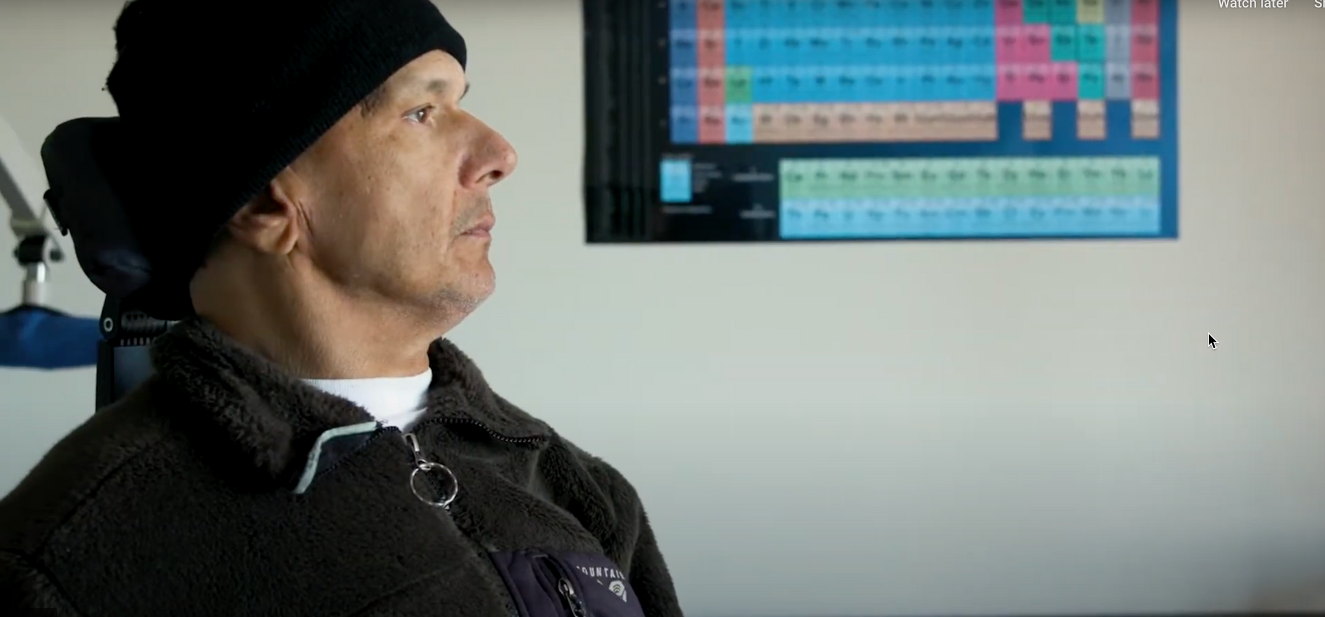 A man wearing a black hat and a black jacket is sitting in front of a periodic table.