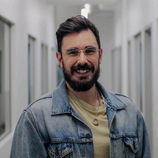 Un homme avec une barbe et des lunettes sourit dans le couloir, portant une veste en jean et une chemise légère.