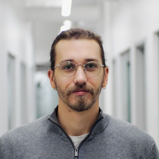 Homme avec des lunettes et une barbe, en pull gris, debout dans un couloir avec des portes blanches.